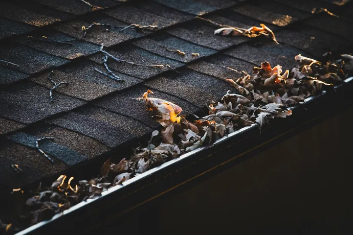 Close-up of a dark shingled roof with a gutter filled with dry leaves and a small flame