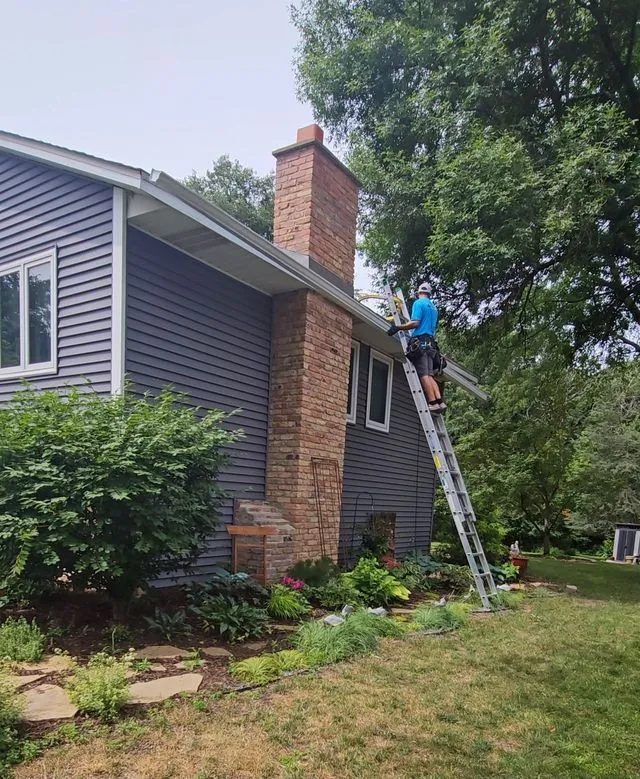 Person on a ladder repairing a house roof beside a brick chimney.