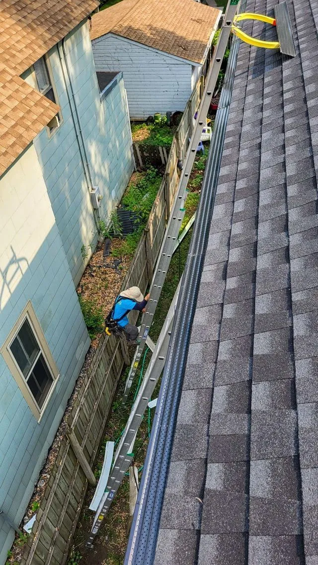 Narrow alley between houses with a blue tarp and ladders beside a gray shingled roof.