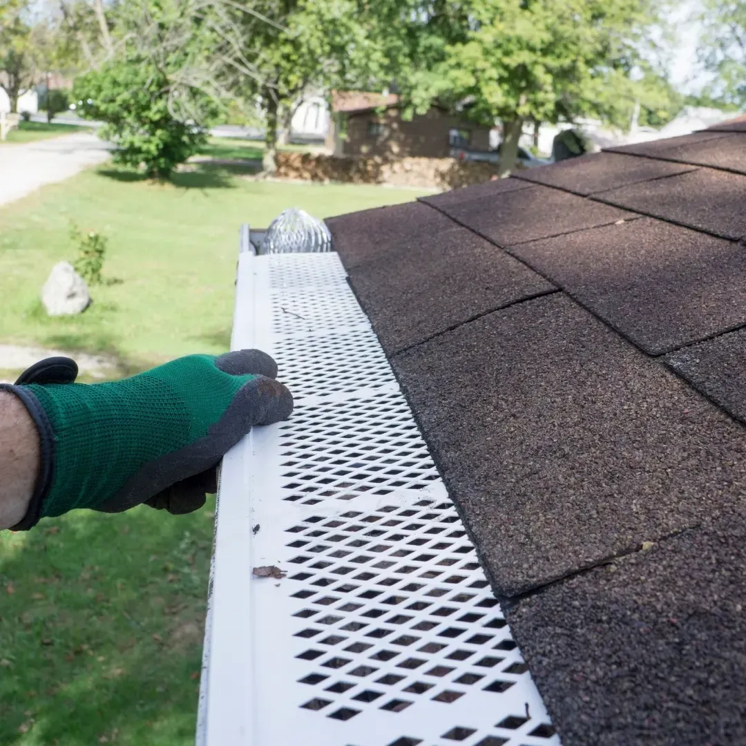 Gloved hand placing white gutter guard along a shingled roof edge outside
