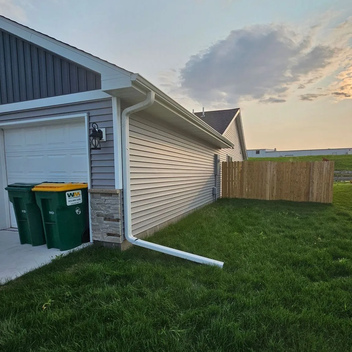 Suburban house side yard with garage, green bins, white siding, and a wooden fence at sunset