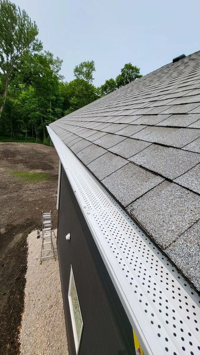 Gutter along a shingled roof edge beside trees and a dirt yard