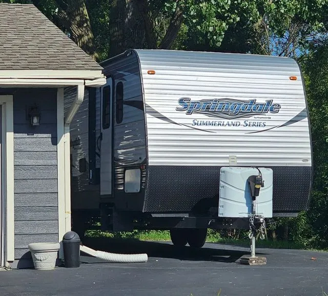 White and gray Spree travel trailer parked beside a house with a white utility hookup and black driveway.
