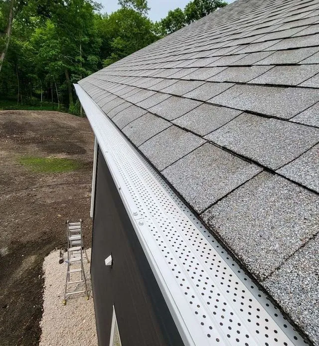 Gray shingled roof with white perforated gutter, viewed along the eave beside trees and bare ground